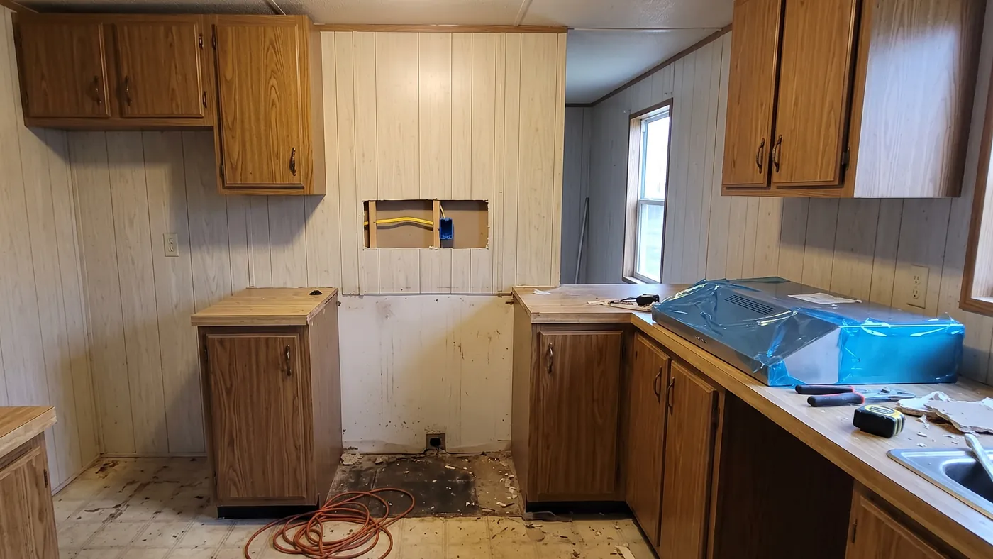A paneled manufactured home kitchen with the range removed, a cut in the wall showing a new outlet box and cable, and a range hood in blue film on the counter